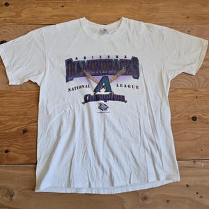 May include: White t-shirt featuring the Arizona Diamondbacks logo in purple, teal, and gold. The design includes the words "Diamondbacks", "National League", and "Champions". Two crossed baseball bats are also part of the design.