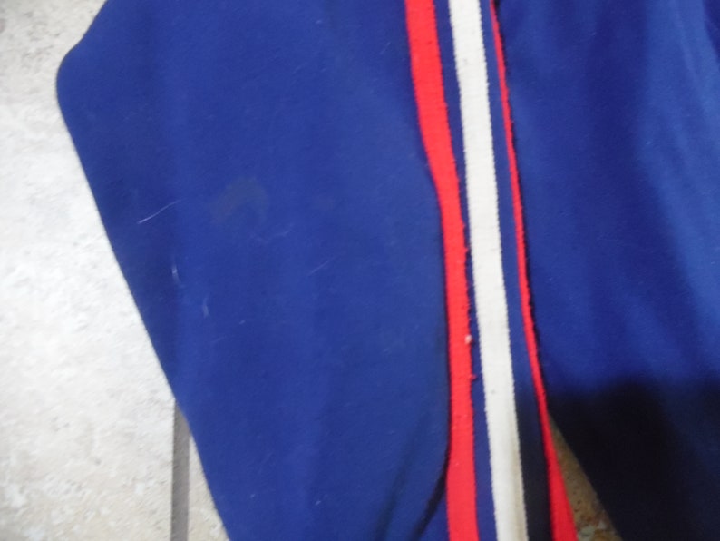 May include: A royal blue garment featuring a vertical side stripe detail. The stripe consists of a central white band flanked by two thin red lines. The fabric appears to be a smooth, solid-colored material with a consistent texture throughout the piece.
