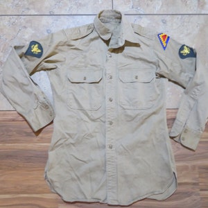 May include: Tan button-down shirt with two chest pockets and patches on the sleeves. The patches feature a yellow eagle and a blue, yellow, and red triangle. The shirt has long sleeves and a collared neckline.