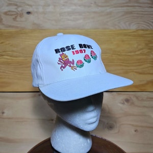 May include: A white baseball cap featuring an embroidered design on the front. The text reads ROSE BOWL 1997 in black and red. Below the text is a purple devil figure holding a pitchfork, followed by three red roses with green leaves.