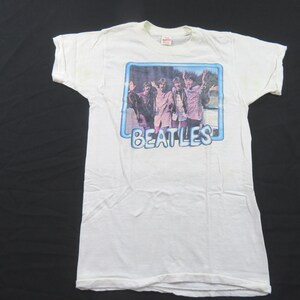 May include: White t-shirt with a graphic of The Beatles within a blue-bordered rectangle. The band members are in a group pose with their arms raised. The word "BEATLES" is printed in blue below the image.