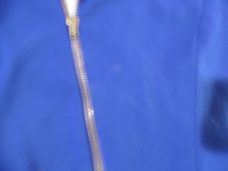 May include: A vibrant royal blue fabric surface featuring a vertical, semi-transparent, textured plastic zipper track running down the center. The zipper pull is a clear, molded plastic component attached to the top of the flexible, ribbed track.