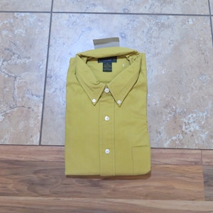 Può includere: Camicia abbottonata giallo senape piegata con colletto button-down e taschino sul petto. La camicia ha bottoni bianchi e un'etichetta in alto. La camicia è realizzata in un tessuto morbido e tessuto.