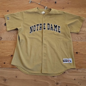 Può includere: Una maglia da baseball dorata con lettere nere che recitano "Notre Dame". La maglia ha una chiusura con bottoni sul davanti e un logo Majestic Apparel sulla manica destra.