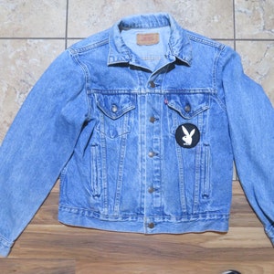 May include: Light blue denim jacket with a classic collar, button closure, and two chest pockets. Features a black and white Playboy patch on the front. A vintage style jacket.