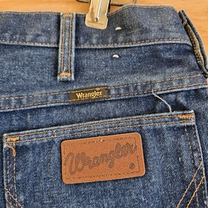 May include: Close-up of the back of blue denim Wrangler jeans. The jeans have a brown leather patch with the Wrangler logo on the back pocket. The stitching is a contrasting gold color. A metal clip is attached to the waistband.