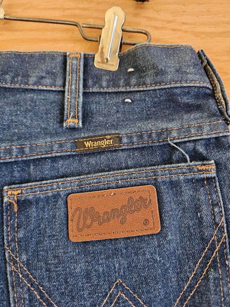 May include: Close-up of the back of blue denim Wrangler jeans. The jeans have a brown leather patch with the Wrangler logo on the back pocket. The stitching is a contrasting gold color. A metal clip is attached to the waistband.