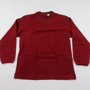 May include: A plain red long-sleeved top. The garment features a crew neck and a straightforward design. The sleeves extend to the wrists. The top is made from a soft, comfortable fabric.