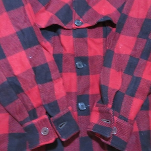 May include: A long-sleeve button-down shirt featuring a bold red and black buffalo plaid pattern. The garment includes a front button placket with dark buttons and matching buttoned cuffs at the end of each sleeve, showcasing a classic checkered design.