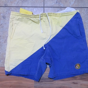 Op de afbeelding: Geel en blauw diagonaal kleurblok shorts met een tailleband met trekkoord. De shorts hebben twee zakken en een klein geborduurd embleem op het blauwe gedeelte. De shorts zijn gemaakt van een lichtgewicht stof.