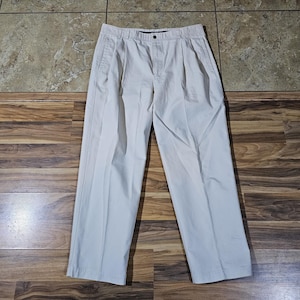 May include: Light beige pleated trousers. The trousers feature a button and zip fastening, belt loops, and two front pockets. They are made of a lightweight fabric and are laid flat on a wooden surface.