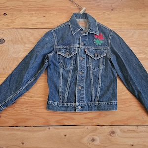 May include: A blue denim jacket with a red and green rose patch on the left chest. The jacket has a button-up front, two chest pockets, and long sleeves. The jacket is displayed on a wooden surface.