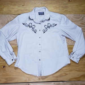 May include: A white long-sleeve button-down shirt with black embroidery of a floral design. The shirt has a pointed collar and a single button closure at the cuff.