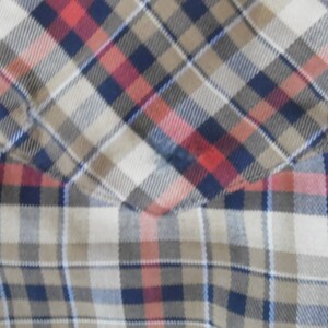 May include: A plaid shirt featuring a chest pocket with a diagonal pattern alignment. The fabric displays a woven check design in shades of navy blue, tan, white, and muted red, creating a consistent grid pattern across the garment surface.