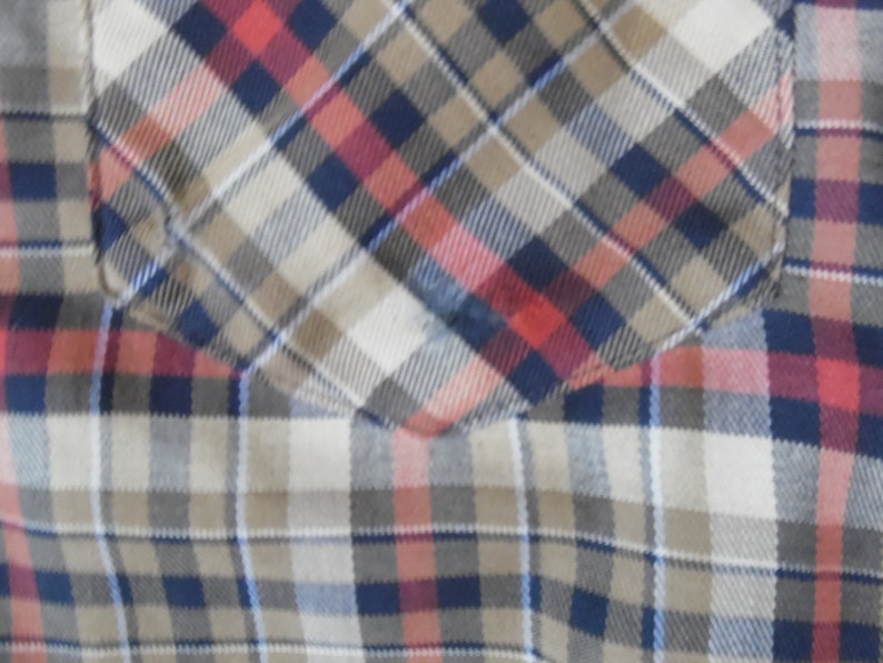 May include: A plaid shirt featuring a chest pocket with a diagonal pattern alignment. The fabric displays a woven check design in shades of navy blue, tan, white, and muted red, creating a consistent grid pattern across the garment surface.