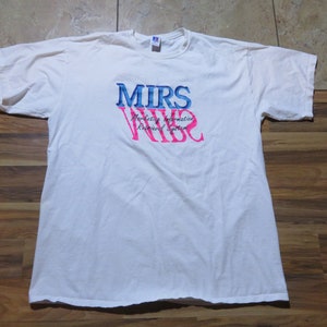 Puede incluir: Camiseta blanca con el texto "MIRS VIBE" en azul y rosa. Las palabras "Marketing Information Retrieval System" están impresas debajo del texto principal. La camiseta está hecha de un material suave y cómodo.