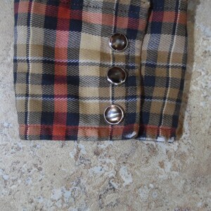 May include: A long-sleeve shirt cuff featuring a plaid pattern in shades of tan, black, and orange. The cuff is secured with three round, dark brown buttons with metallic silver rims, arranged in a vertical line along the side of the fabric.