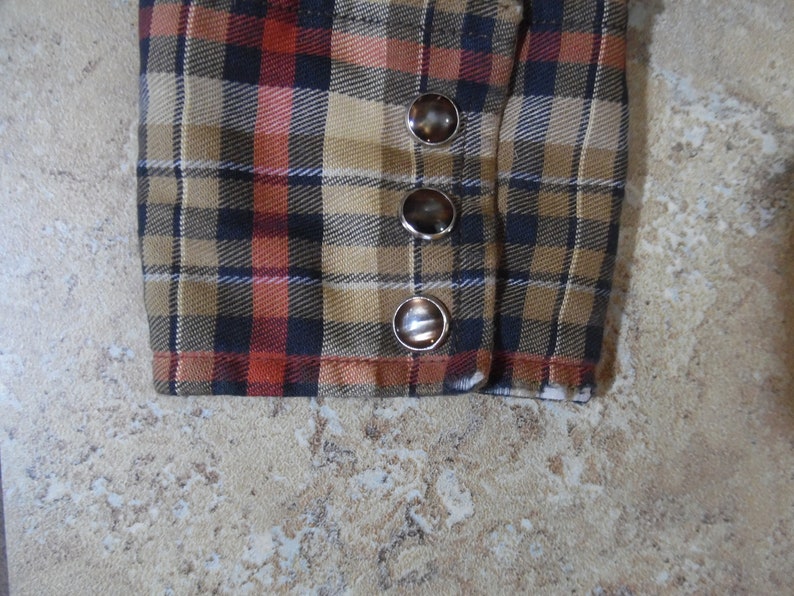 May include: A long-sleeve shirt cuff featuring a plaid pattern in shades of tan, black, and orange. The cuff is secured with three round, dark brown buttons with metallic silver rims, arranged in a vertical line along the side of the fabric.