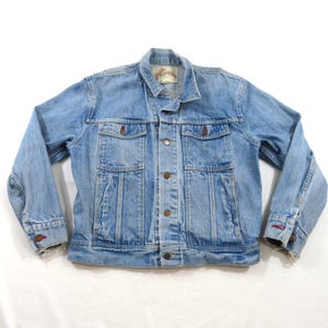 May include: A light blue denim jacket featuring a classic collar, a button-up front, and two chest pockets with buttoned flaps. The jacket has long sleeves with buttoned cuffs, metal hardware, and a visible interior label at the neckline.