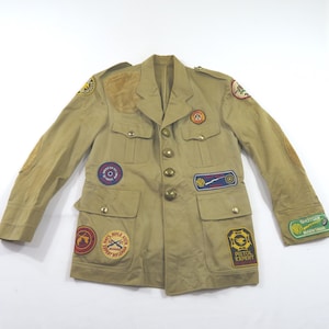 May include: A tan long-sleeve jacket featuring multiple embroidered patches, including NRA-themed insignia, a shoulder patch, and elbow patches. The jacket has a button-down front, two chest pockets, two lower pockets, and a collar with epaulets.