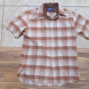 May include: Short-sleeved button-up shirt with a plaid pattern in shades of brown and white. The shirt has a quilted brown collar and a single chest pocket. The fabric appears to be a soft, woven material.