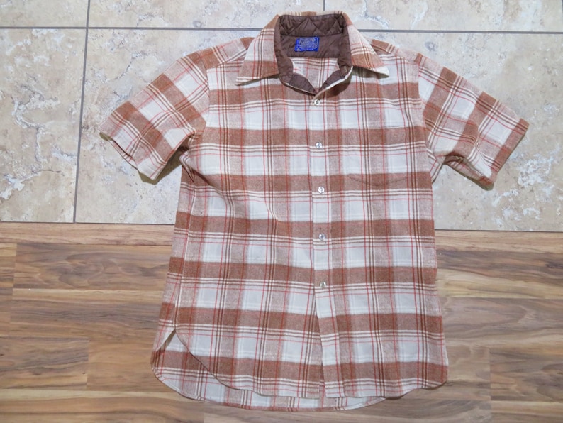 May include: Short-sleeved button-up shirt with a plaid pattern in shades of brown and white. The shirt has a quilted brown collar and a single chest pocket. The fabric appears to be a soft, woven material.