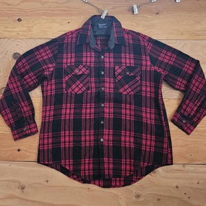 Pode incluir: Uma camisa de flanela xadrez vermelha e preta com dois bolsos no peito e uma frente abotoada. A camisa tem gola e mangas compridas. O tecido parece ser de peso médio, adequado para climas mais frios.