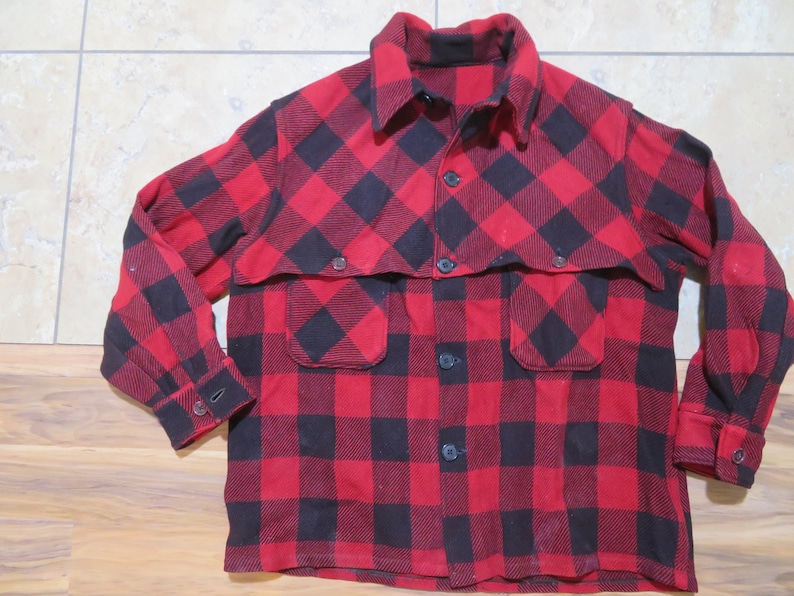 May include: A long-sleeve button-up shirt featuring a red and black buffalo plaid pattern. The shirt includes a pointed collar, a front button closure, two chest pockets with flaps, and buttoned cuffs on the sleeves. The fabric has a textured finish.