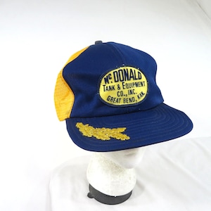 Op de afbeelding: Een blauwe en gele truckerpet met een gele ovale patch met de tekst "MC DONALD TANK & EQUIPMENT CO., INC. GREAT BEND, KAN." in het blauw. De pet heeft een goudkleurig geborduurd bladmotief aan de linkerkant van de donkerblauwe klep.