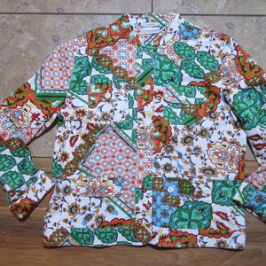 Op de afbeelding: Een shirt met lange mouwen en een patchwork-ontwerp. Het shirt heeft een witte achtergrond met kleurrijke bloemen- en geometrische patronen in oranje, groen, blauw en bruin. Het heeft twee voorzakken.