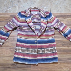 May include: A colorful blazer with a Southwestern-inspired pattern. The jacket features a button closure, two front pockets, and a notched collar. The design includes stripes in shades of blue, red, brown, and cream, with geometric accents.
