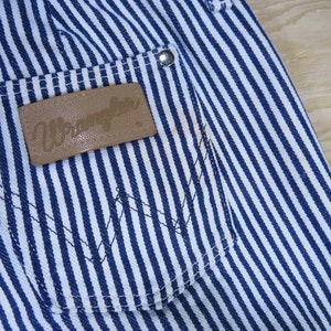 May include: Blue and white striped denim pants with a brown leather patch that says "Wrangler" on the back pocket.