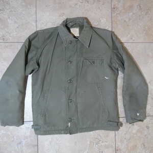 May include: Olive green work jacket with a button-up front, two chest pockets, and two side pockets. The jacket has a collar and adjustable straps at the waist. The sleeves are long and the jacket appears to be lightly insulated.