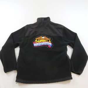 May include: A black fleece jacket featuring a high collar and long sleeves. The back displays a large embroidered patch of an eagle with spread wings above a blue and red banner containing the white text Harley-Davidson Motorcycles.