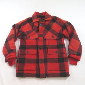 May include: A red and black buffalo plaid jacket featuring a pointed collar, a front button closure, and four front pockets with buttoned flaps. The long sleeves have ribbed cuffs, and the garment is constructed with a thick, textured fabric.
