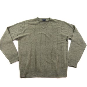 Könnte beinhalten: Ein langärmeliger V-Ausschnitt-Pullover in einem gedeckten Salbeigrün. Das Strickgewebe weist ein dezentes vertikales Zopfmuster auf der Vorderseite auf. Das Kleidungsstück hat einen gerippten Saum und gerippte Bündchen sowie ein kleines dunkles Etikett am Halsausschnitt.