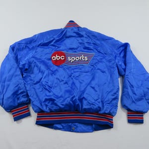 May include: A bright blue satin bomber jacket featuring a red, white, and blue striped collar, cuffs, and waistband. The back displays an embroidered patch with the text abc sports in white and red lettering on a blue and red background.