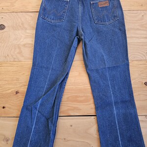 May include: Blue denim jeans with a classic five-pocket design. The jeans feature a brown leather patch with the "Wrangler" logo on the back pocket. The jeans are hanging on a metal hanger against a wooden background.