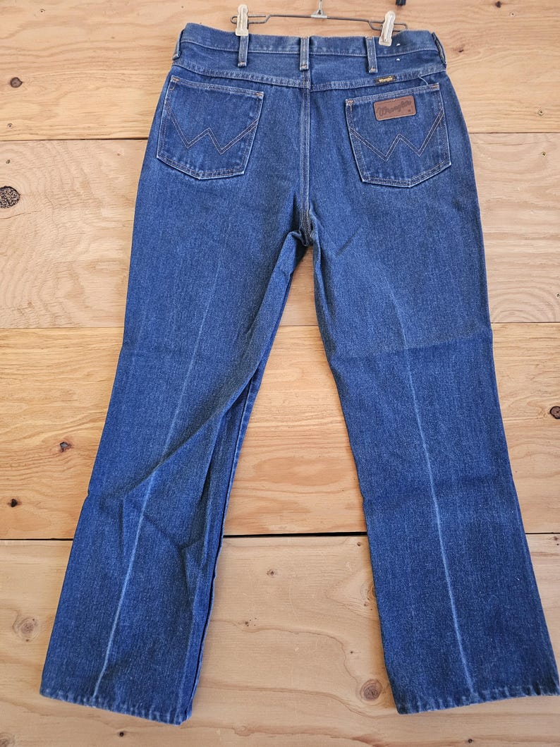 May include: Blue denim jeans with a classic five-pocket design. The jeans feature a brown leather patch with the "Wrangler" logo on the back pocket. The jeans are hanging on a metal hanger against a wooden background.