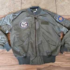 Peut inclure: Veste de bombardier vert olive avec fermeture éclair, poignets côtelés et deux poches avant. Comprend des écussons sur la poitrine et la manche, dont un écusson circulaire avec un avion et le texte "B-17 Flying Fortress".