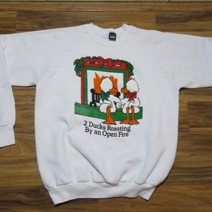 May include: White sweatshirt with a Christmas-themed graphic of two ducks roasting by an open fire. The graphic features a fireplace with a fire, a green wreath, and a red bow. The text "2 Ducks Roasting By an Open Fire" is printed below the graphic.