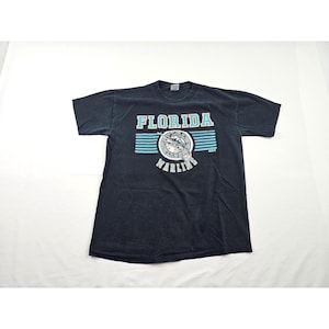 Könnte beinhalten: Ein schwarzes Kurzarm-T-Shirt mit dem Schriftzug FLORIDA MARLINS in hellblauen und weißen Buchstaben. Die Grafik enthält ein kreisförmiges Logo mit einer Marlin-Fisch-Illustration und horizontale hellblaue Streifen im Brustbereich.