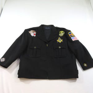 May include: A black jacket featuring a front zipper, two chest pockets with flaps, and various embroidered patches including a tiger head, a masonic square and compass symbol, a military emblem, a United States flag, and a cross on the sleeve.