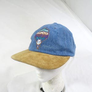 May include: A blue denim baseball cap with a tan suede brim. The front features an embroidered logo with the text 'INAUGURAL SEASON' and 'DIAMOND BACKS' above a graphic of a snake head and a baseball, with the word 'DIAMONDBACKS' stitched below.