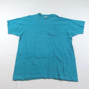 Könnte beinhalten: Ein türkisblaues Kurzarm-T-Shirt mit Brusttasche. Das Shirt besteht aus einem weichen Material und liegt flach auf einer weißen Oberfläche. Das T-Shirt hat einen Rundhalsausschnitt und eine lockere Passform.