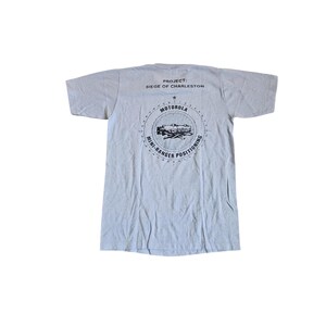 以下が含まれることがあります： 背面に黒いグラフィックがプリントされたライトグレーの半袖Tシャツ。テキストには「PROJECT: SIEGE OF CHARLESTON」および「MOTOROLA MINI-RANGER POSITIONING」と記されており、円形のコンパスの中に技術機器のイラストが中央に配置されています。