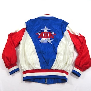 May include: A red, white, and blue satin bomber jacket featuring a large graphic on the back with the text ABA centered inside a stylized white star. The jacket has long red sleeves, a blue center panel, white side panels, and a striped ribbed hem.
