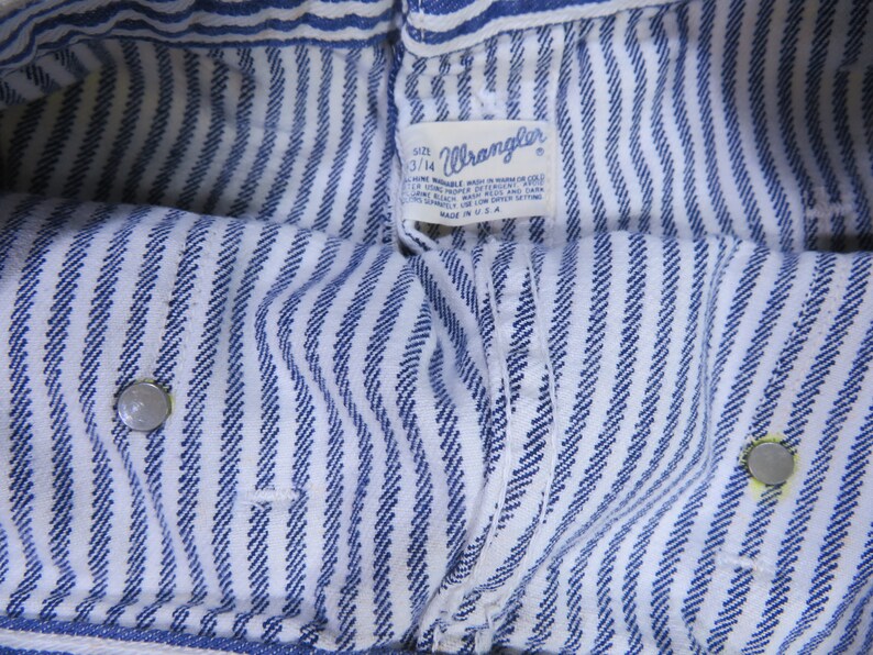 May include: Close-up of blue and white striped fabric with a Wrangler label. The label reads "Size 33/14 Machine Wash in Warm or Cold Water.  Do Not Bleach.  Wash with Like Colors.  Tumble Dry Low.  Made in U.S.A."
