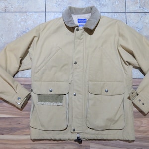 Op de afbeelding: Een beige jas met een corduroy kraag en meerdere zakken. De jas heeft een knoopsluiting aan de voorkant, drukkers op de zakken en een trekkoord aan de onderkant. De mouwen hebben manchetten met knopen.