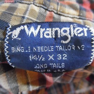 May include: A dark blue Wrangler shirt label with white text reading Wrangler, Single Needle Tailoring, 14 1/2 x 32, and Long Tails, stitched onto a plaid fabric with brown, orange, and black tones. The label also indicates Made in U.S.A.